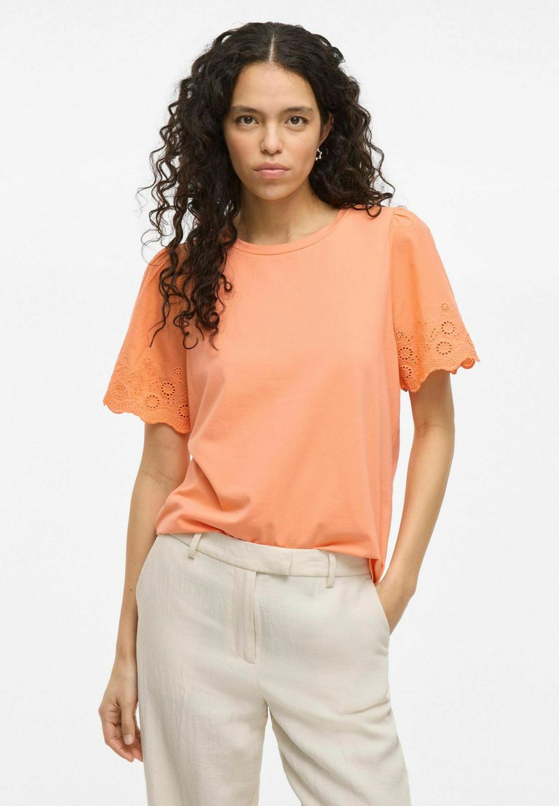 Top de algodón suave de color coral con mangas cortas y detalles de encaje cortado con láser en las mangas, combinado con pantalones de corte crema.
