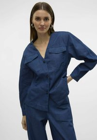 Giacca di jeans blu scuro con scollo a V, maniche lunghe, due tasche frontali e chiusura con bottoni. Texture liscia e design strutturato.