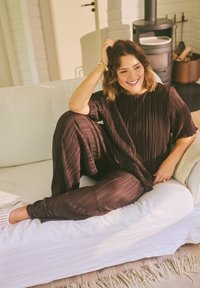 Femme aux cheveux ondulés portant une tenue plissée marron, assise en souriant sur un canapé blanc dans une pièce chaleureuse et ensoleillée avec un poêle à bois.