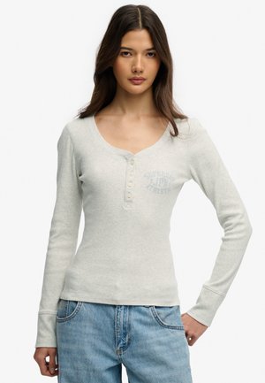 Młoda kobieta o długich ciemnych włosach, ubrana w jasnoszarą, długorękawową, zapinaną na guziki bluzkę oraz jasnoniebieskie dżinsy, stoi z jedną ręką na biodrze.