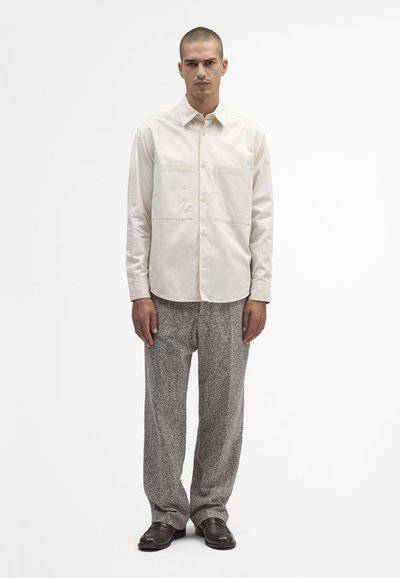 Beige skjorte med knapper og to forlommer, parret med strukturerede grå bukser og brune sko på en ensfarvet hvid baggrund.