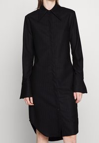 Svart randig skjortklänning med spetsig krage, långa ärmar och utsvängda manschetter. Har knappslå framtill och sidofickor.