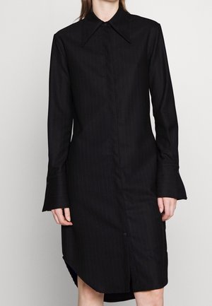 Abito camicia a righe nere con colletto a punta, maniche lunghe e polsini svasati. Presenta una chiusura frontale con bottoni e fessure laterali.