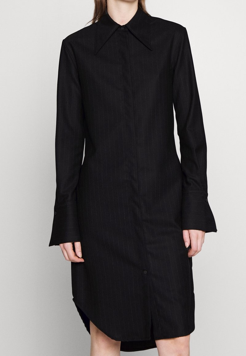 Svart randig skjortklänning med spetsig krage, långa ärmar och utsvängda manschetter. Har knappslå framtill och sidofickor.