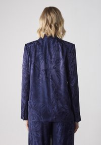 Tumman sininen jacquard-pukukoira, jossa on kukkakuosi. Se sisältää muotoiltua leikkausta, notkeat kaulukset ja napitettu hihansuu, näkyy takaa.