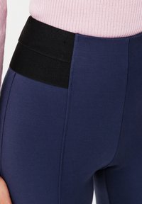 Marineblauwe leggings met een gladde textuur en een brede zwarte elastische tailleband. Beschikt over een zijnaaddetail voor een extra ontwerpelement.
