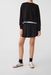 Zwarte cropped sweatshirt met wijde mouwen, grijs gelaagd shirt eronder, zwarte plissé skort en witte sneakers met zwarte accenten.