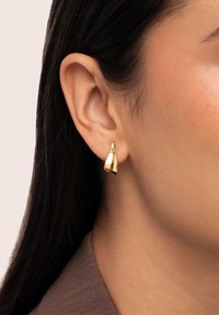 Goldfarbener, tropfenförmiger Ohrring mit glänzender Oberfläche, der am Ohrläppchen befestigt ist und seine glatte Textur sowie modernes Design betont.