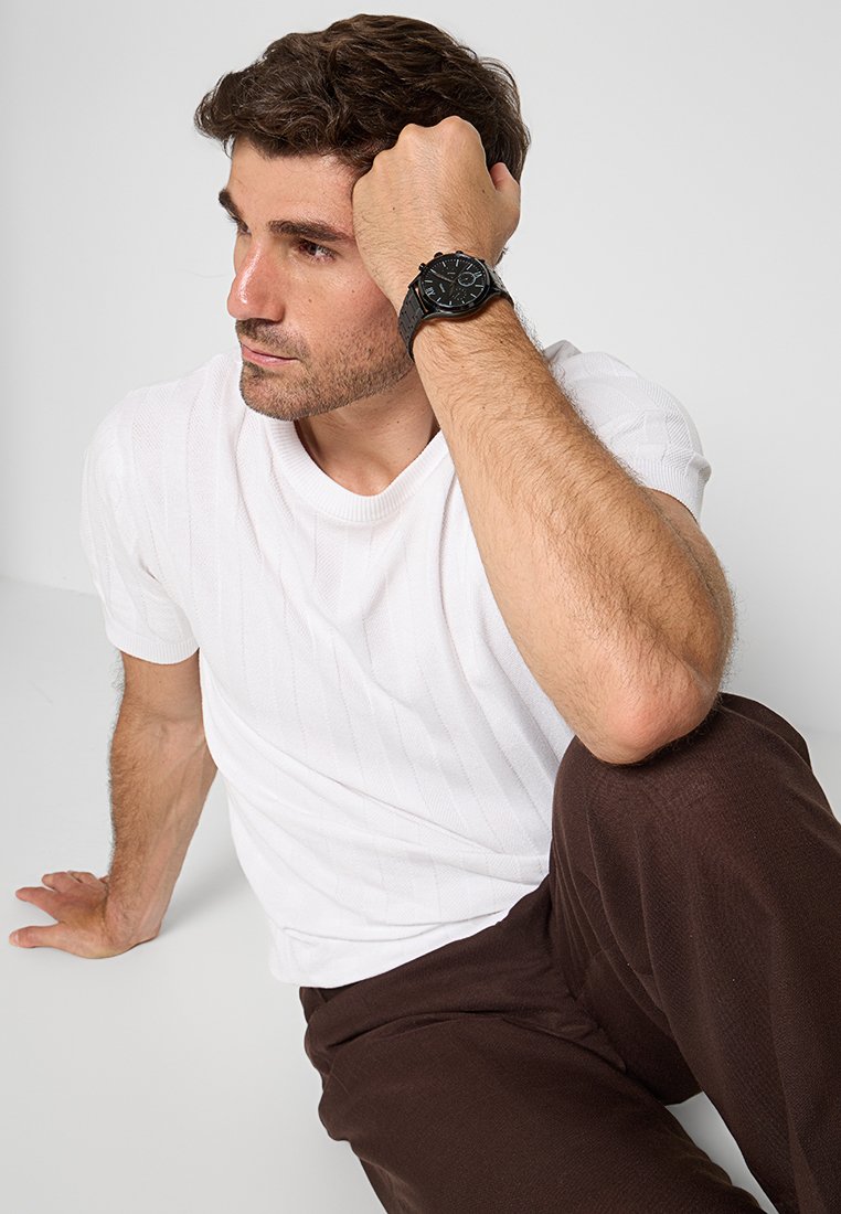 Schwarze Armbanduhr, weißes strukturiertes Strick-T-Shirt, braune Hose. Die Uhr hat ein rundes Ziffernblatt mit mehreren Ziffern und ein Lederarmband.