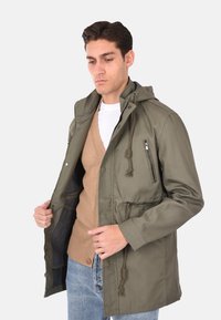 Giacca leggera verde oliva con cappuccio, chiusura con zip, vita regolabile con cordino e tasche laterali, indossata sopra un maglione beige.