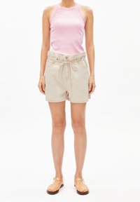 Mujer vestida con una blusa sin mangas rosa, pantalones cortos de tiro alto beige con un lazo, y sandalias translúcidas naranjas, de pie contra un fondo blanco.