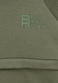 Zöld szövet, amely puha tapintású; tartalmaz egy struktúrált panelt és egy dombornyomott "Blue Ridge Denim" logót, amely megegyező színű.