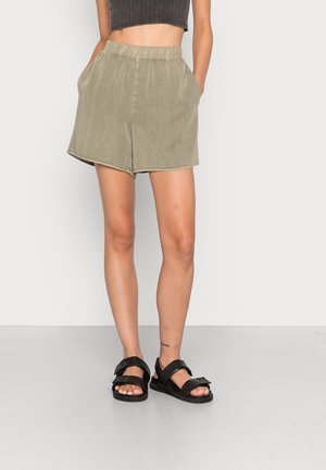 Personne portant un short à taille élastique vert olive et des sandales noires, debout les mains dans les poches devant un fond clair uni.
