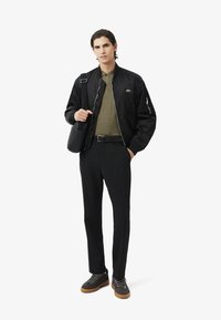 Chaqueta bomber negra, camisa verde oliva, pantalones negros, zapatos grises con suelas marrones. Modelo sosteniendo una bolsa de hombro negra, de diseño minimalista.