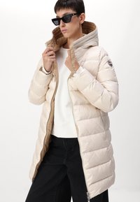 Beige gewatteerde pufferjas met een capuchon, voorzien van een contrasterende bruine binnenvoering en ritssluiting. Draagt over een witte trui en zwarte broek.