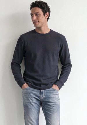 Homme aux cheveux bouclés portant une chemise sombre à manches longues et un jean bleu clair, debout les mains dans les poches devant un fond uni.
