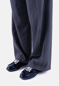 Zapatos azul marino con una combinación de materiales brillantes y texturizados, con cordones blancos y un diseño de punta único, combinados con pantalones de pierna ancha grises.