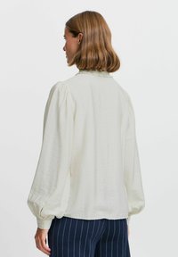 Cremefarbene Bluse mit langen Puffärmeln, rundem Kragen und geknöpften Manschetten, kombiniert mit navyblauen Nadelstreifen-Hosen. Leichtes, strukturiertes Material.