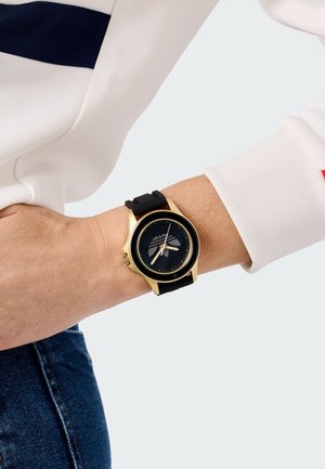 Montre avec bracelet en silicone noir, boîtier en métal doré, cadran noir avec des accents blancs et un design graphique épuré sur le visage.