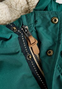 Veste vert foncé avec doublure en Sherpa, dotée d'une fermeture éclair, d'une tirette en cuir et de boutons-pression en laiton. Conception matelassée avec couture contrastée.