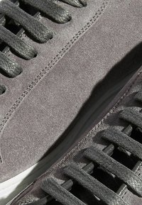 Graue Wildleder-Sneaker mit strukturiertem Finish, ausgestattet mit schwarzen flachen Schnürsenkeln und einer weißen Gummisohle. Eine Nahaufnahme hebt die Nähdetails hervor.