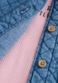 Gevoerde denim buitenlaag in een lichtblauwe tint, die over een geribbeld roze kledingstuk valt. Bevat een beige knop en subtiele stikdetails.