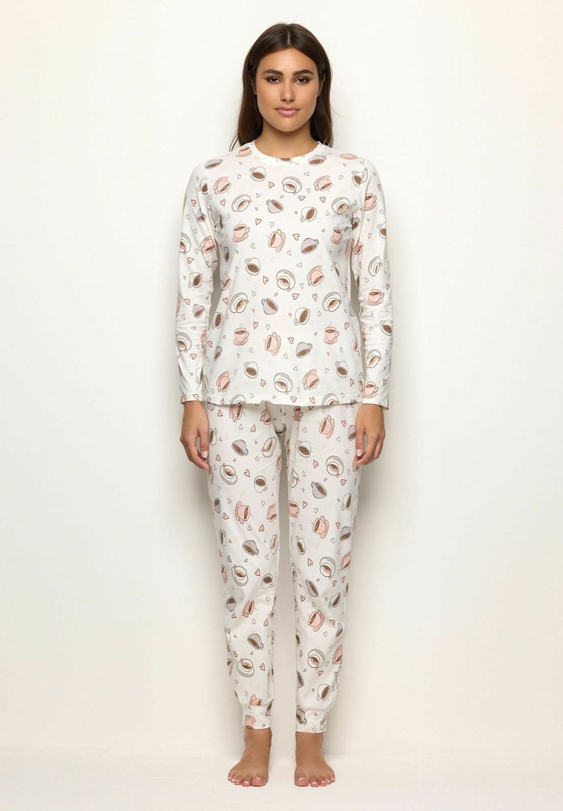 Junge Frau steht barfuß in einem weißen langärmeligen Pyjama-Set mit braunen und rosa Kaffeetassen- und Herzmustern.