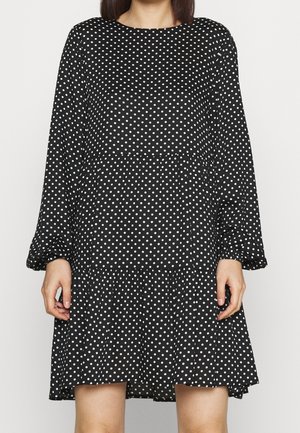 Frau trägt ein schwarzes Langarmkleid mit weißen Polka-Dots, steht vor einem schlichten hellen Hintergrund.
