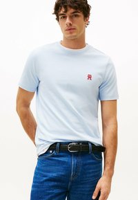 T-shirt di cotone azzurro chiaro con maniche corte e colletto rotondo, caratterizzata da un piccolo logo rosso sul petto. Abbinata a jeans in denim scuri e una cintura nera.