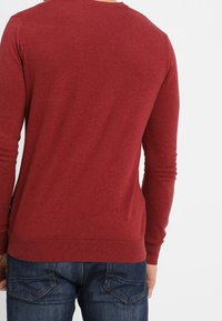 Czerwony sweter z długim rękawem, wykonany z dzianiny, z okrągłym dekoltem i prążkowanym wykończeniem na brzegu. Tkanina wydaje się miękka, o gładkiej fakturze. Noszony na niebieskich dżinsach.