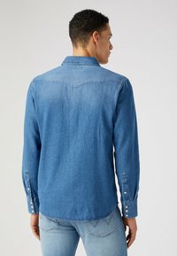 Camisa de mezclilla azul con mangas largas y puños abotonados, que cuenta con un yugo trasero y un dobladillo redondeado. Textura suave con un efecto de lavado ligero.
