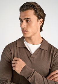 Ruskea teksturoitu polo-paita, jossa on kauluspaita, valkoisen pyöriäispaitojen päällä, mallin käsi ristissä rinnan päällä.