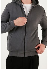 Felpa grigia con zip, dotata di cappuccio foderato in fleece testurizzato, tasche frontali e vestibilità comoda. Presenta polsini e cinturino a coste, oltre a un tessuto liscio.