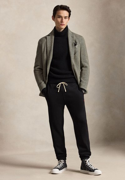 Giovane uomo in piedi con le mani nelle tasche che indossa un blazer a spina di pesce, un dolcevita nero, pantaloni da jogging neri con coulisse bianca e sneakers nere alte.