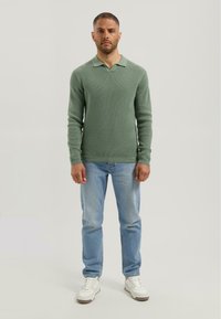 Man standing facing forward wearing a textured green long-sleeve polo shirt, light blue jeans, and white sneakers on a plain background.