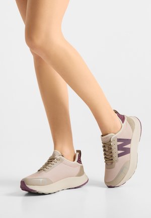 Jambes portant des baskets beiges et violettes à lacets avec des semelles épaisses, posées sur un fond gris clair.