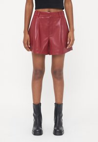 Shorts in pelle bordeaux con texture liscia, pieghe davanti e lunghezza a metà coscia. Abbinati a stivali neri alla caviglia.