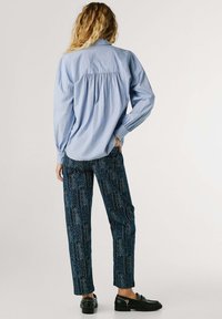 Lichtblauw, langarm overhemd met gather-achterkantdetail, in combinatie met donkerblauwe, gemusterde broek en zwarte instappers.