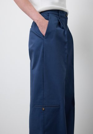 Person trägt marineblaue, maßgeschneiderte Hose mit geknöpftem Taschen-Detail und Hand in Seitentasche, kombiniert mit weißem Oberteil.