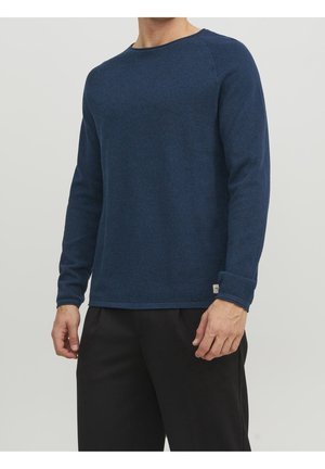 Mann trägt einen langärmligen, strukturierten marineblauen Pullover und schwarze Hose vor einem schlichten hellgrauen Hintergrund.