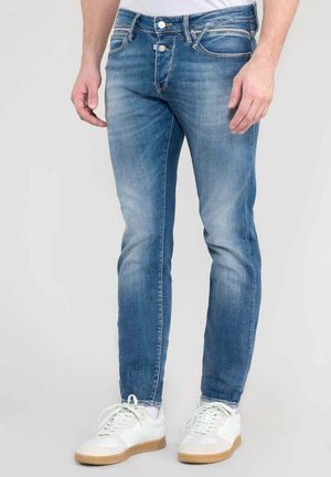 Vaqueros de mezclilla azul de corte ajustado, con un ligero desgaste, bolsillos frontales y cremallera. Llevados con zapatillas blancas y una camiseta básica.