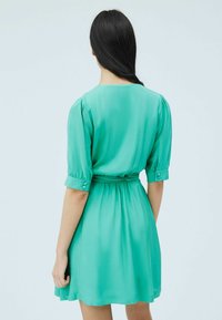 Vihreä satiinimekko, jossa on lyhyet puffihihoja, vyötärösidonta ja nappidetaljeja. Sujuva pinta, polven ylle ulottuva pituus ja hieman levenevä hame.