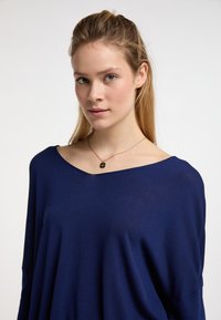 Maglia in maglia blu navy, dal taglio ampio e con scollatura larga e maniche lunghe. Tessuto leggero con texture liscia. Collana con pendente dorato visibile.