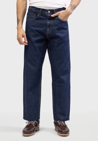 Dark blue straight-leg jeans with a high waist, featuring five pockets and a classic button closure. Worn with brown lace-up shoes.