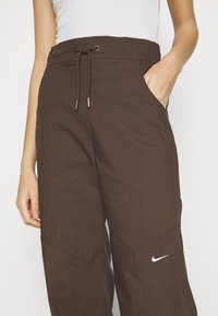 Pantalons marron, coupe ample avec une taille élastique et un cordon de serrage ; dotés de poches latérales et d'un petit logo Nike blanc sur la jambe.