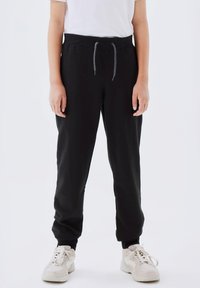 Black sweatpants with an elastic waistband, adjustable drawstring, tapered legs, and ribbed cuffs, paired with white sneakers.