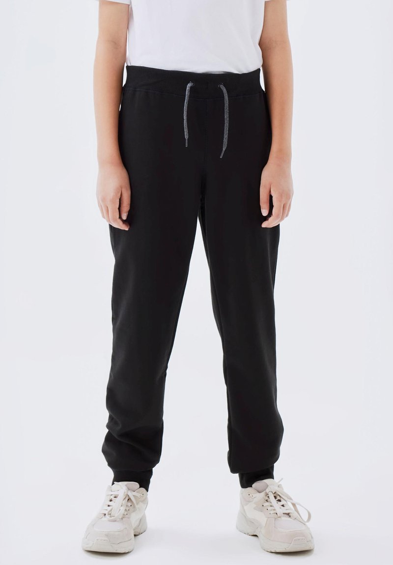 Black sweatpants with an elastic waistband, adjustable drawstring, tapered legs, and ribbed cuffs, paired with white sneakers.