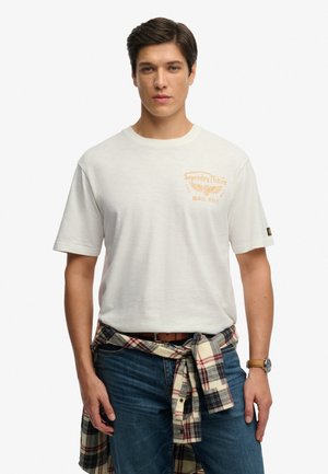 Hombre con camiseta de Superdry Motors color blanco hueso, jeans azules, camisa de cuadros atada alrededor de la cintura y reloj de pulsera, de pie frente a un fondo blanco.