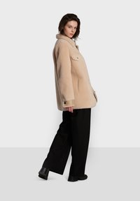 Casaco de pele sintética bege com colarinho em bico, botões frontais e bolsos laterais, combinado com calças largas pretas e botas de salto bloco.