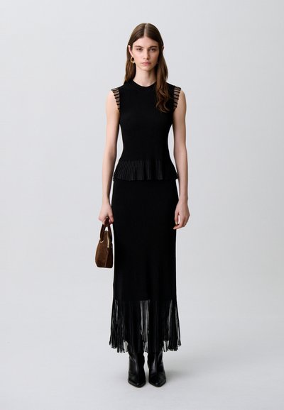 Donna con vestito nero senza maniche con dettagli plissettati in vita e sull'orlo, indossa stivali neri e tiene una piccola borsa marrone contro uno sfondo neutro.
